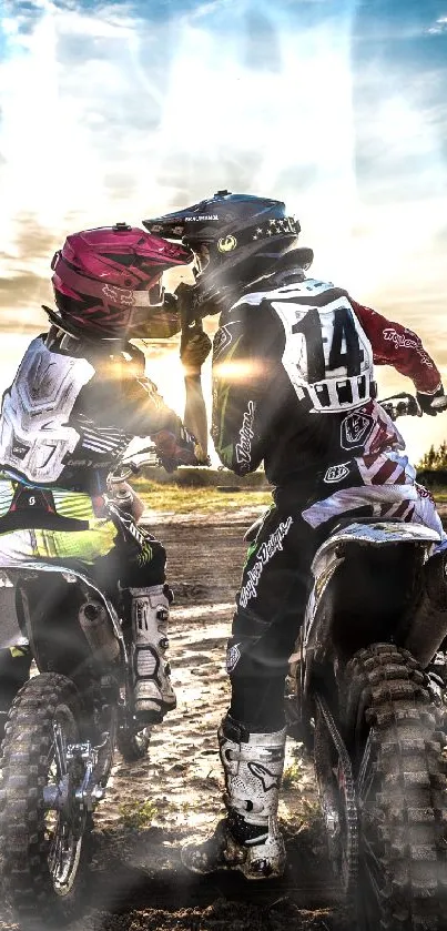 Two motocross riders share a moment at sunset on a dirt track.