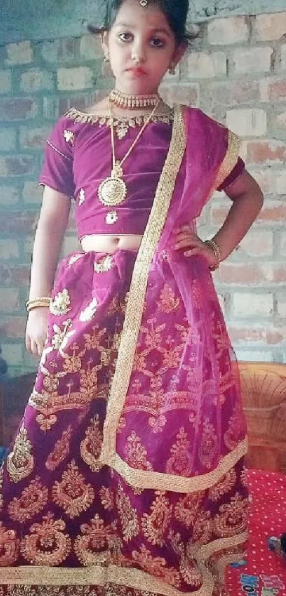 Child in maroon traditional dress against brick wall.