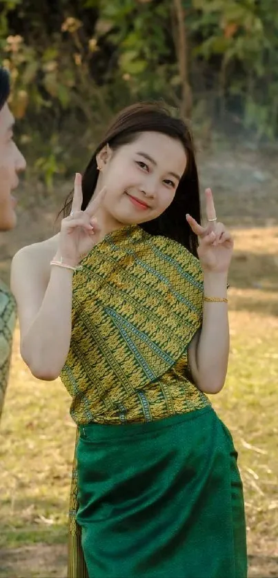 Cheerful woman in green traditional attire makes a peace sign in nature.