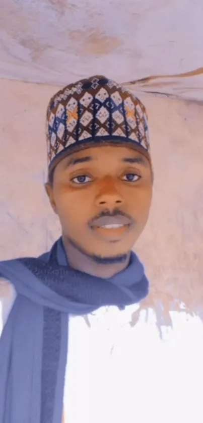 Man in traditional attire with patterned hat against a beige wall.