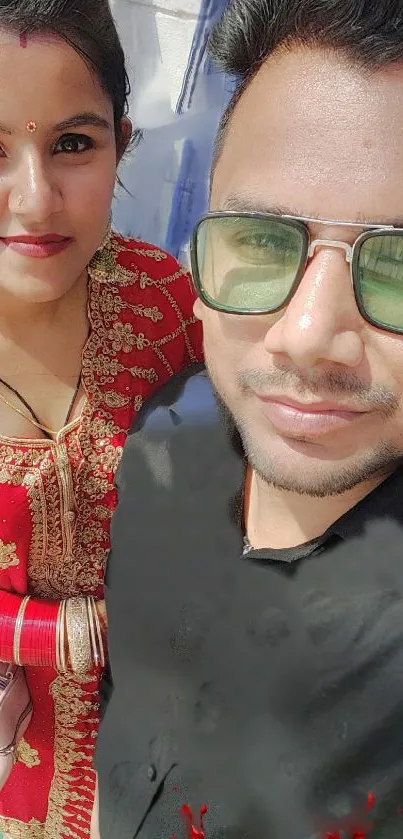 Selfie of couple in traditional red attire with sunglasses, outdoor setting.