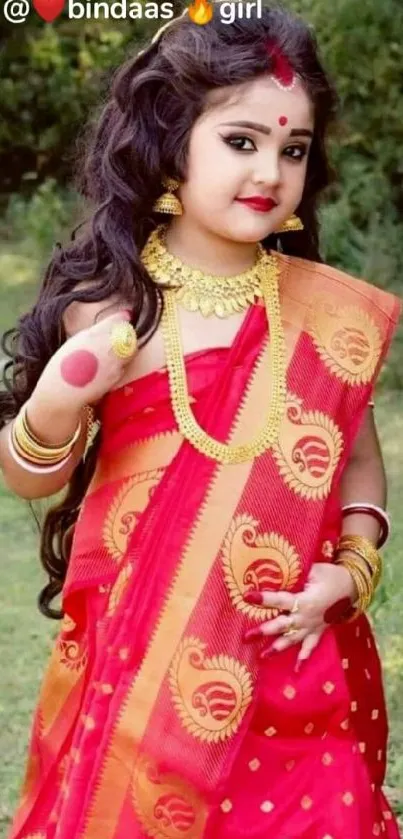 Young girl in traditional red saree with gold jewelry against green backdrop.