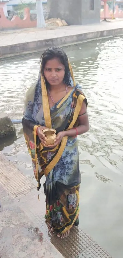 Woman in a vibrant sari by the water with traditional items.