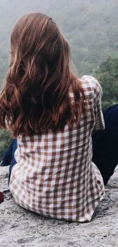 Woman in a plaid shirt sitting on a rock overlooking misty mountains.