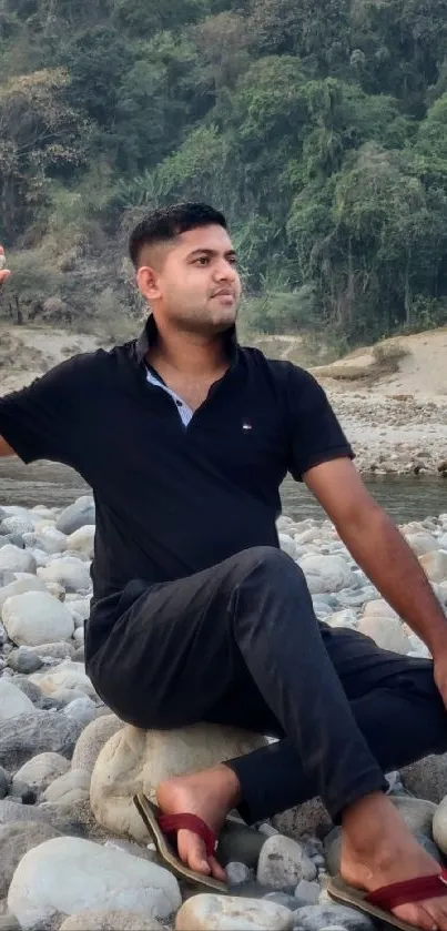 A man sitting peacefully on a rocky riverbank amidst nature.