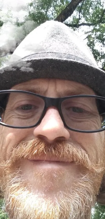 Man wearing hat under green trees, smiling casually.