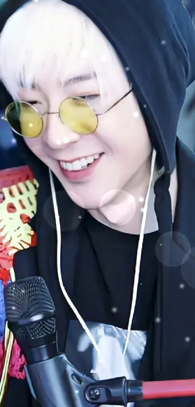 Person wearing a black hoodie and yellow glasses, smiling with a microphone.