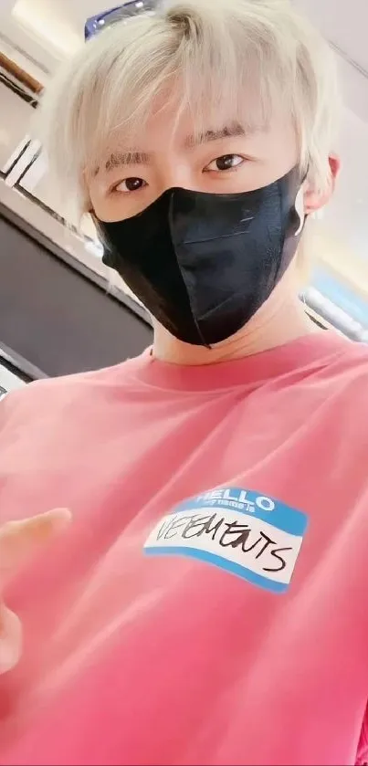 Person wearing pink shirt and black mask, showing peace sign.