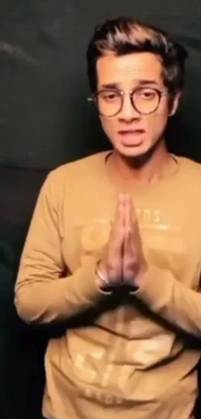 Young man with glasses, hands clasped, dark background.