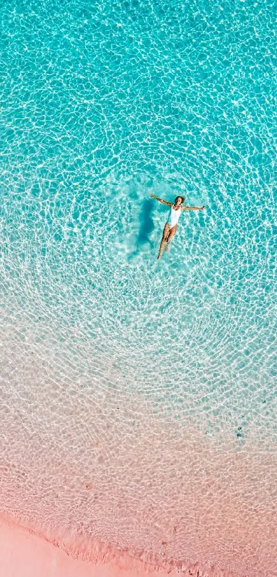 Aerial view of a person floating in turquoise ocean water on pink sand.