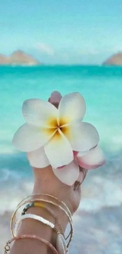 Hand holds white flower over turquoise beach.