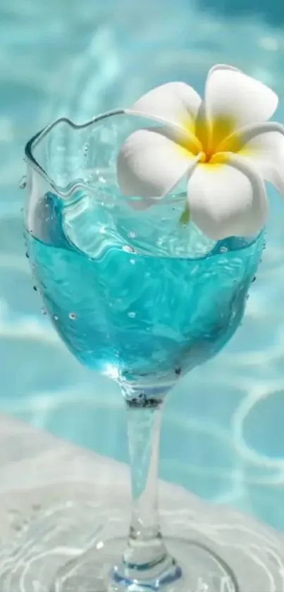 A clear glass with blue water and a white flower by the pool.