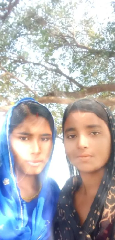 Two women stand under a large tree.