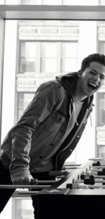 Black and white photo of a man playing foosball and laughing near large windows.