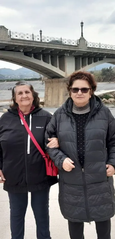 Two individuals walking near an urban bridge with river view.