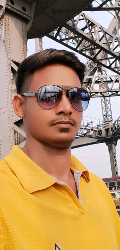 Person wearing sunglasses and a yellow shirt under a metal bridge.