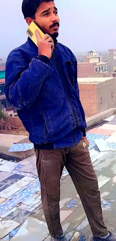 Man in blue jacket on rooftop with phone, vibrant cityscape background.