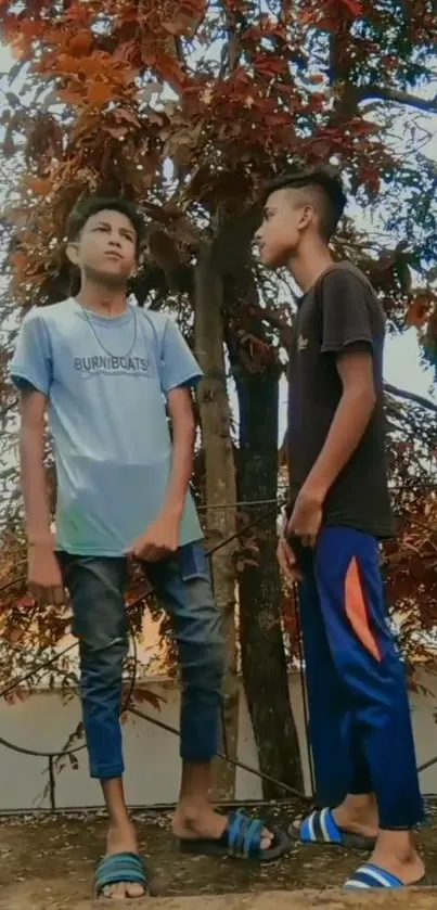 Two individuals in urban attire stand by a tree with autumn leaves.