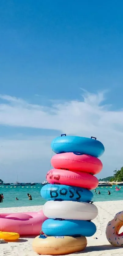 Colorful floaties stacked on a bright beach with a calm ocean and blue sky.