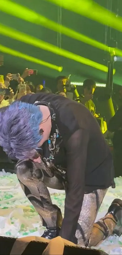 Blue-haired performer kneeling during a vibrant concert with neon lights.