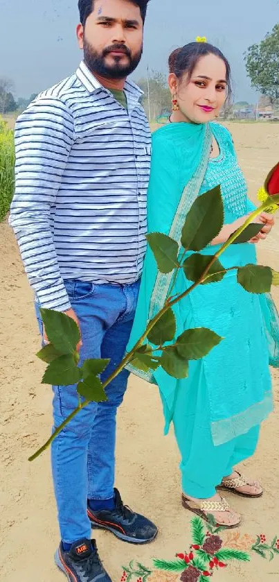 Couple in a rural setting holding a rose with lush fields in the background.