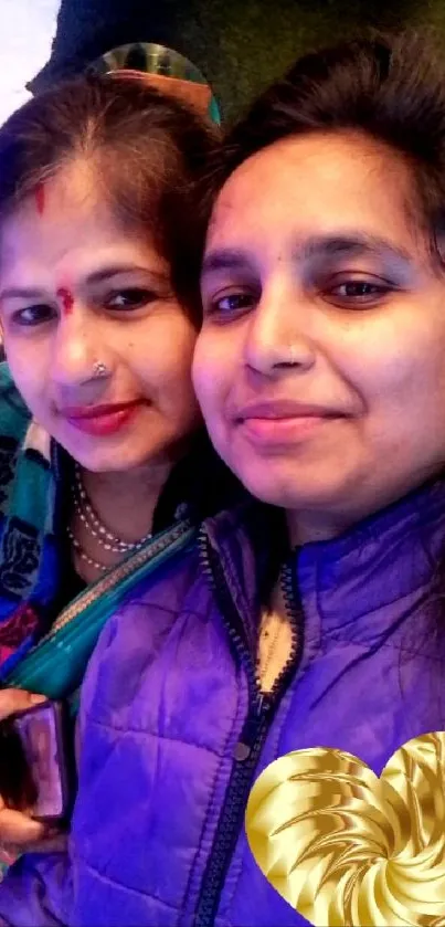 Two smiling women in colorful attire with a golden heart design.