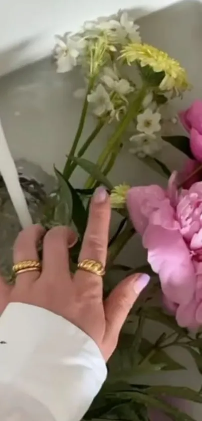 Hand touching pink peonies in water with yellow and white flowers.