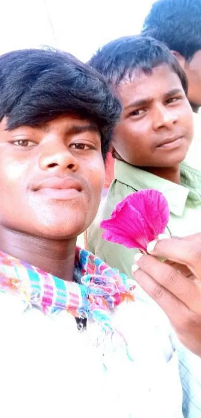 Two friends holding a pink flower, smiling vividly.