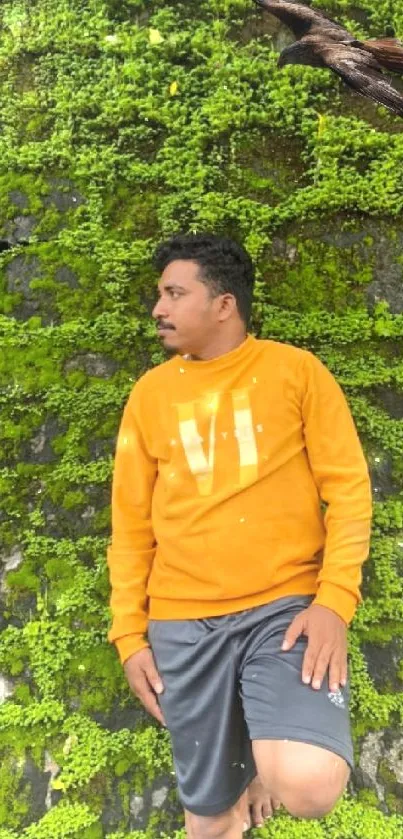 Man stands against a lush green leafy wall wearing an orange shirt.