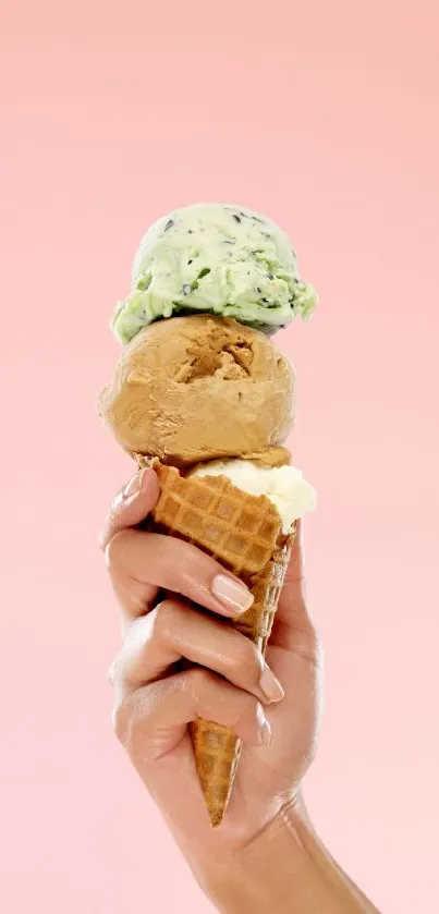 A hand holding a triple scoop ice cream cone against a pink background.