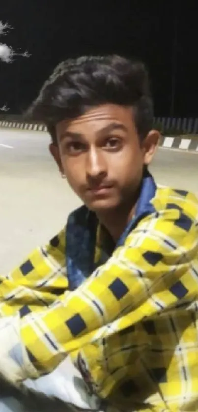 Young man in yellow plaid shirt sitting on a quiet night street.