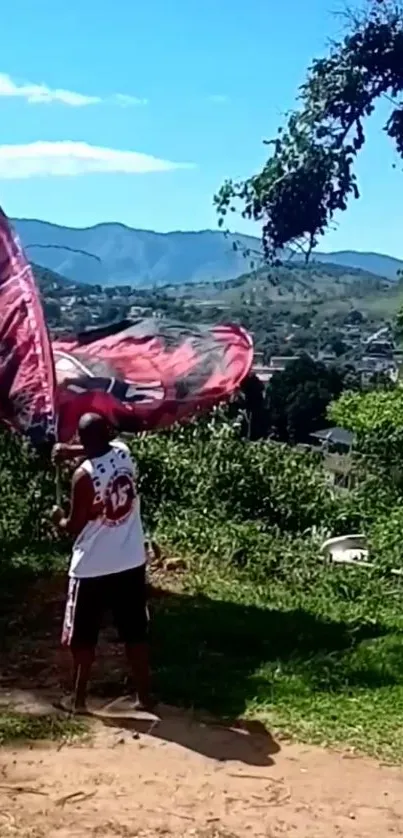 A person waving a large flag on a sunny hillside with mountains in the background.