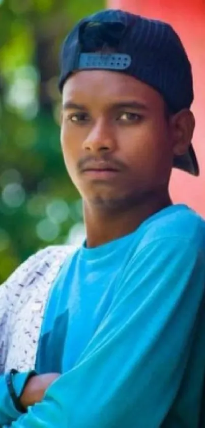 Young man in blue shirt and hat leaning against pink wall with green foliage.