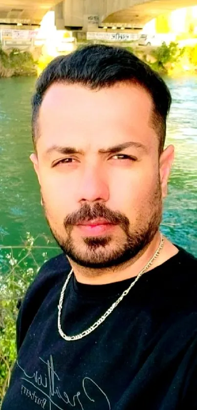 Man standing by a serene river with a bridge in the background.