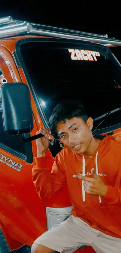 Young man poses next to an orange truck wearing a red jacket.