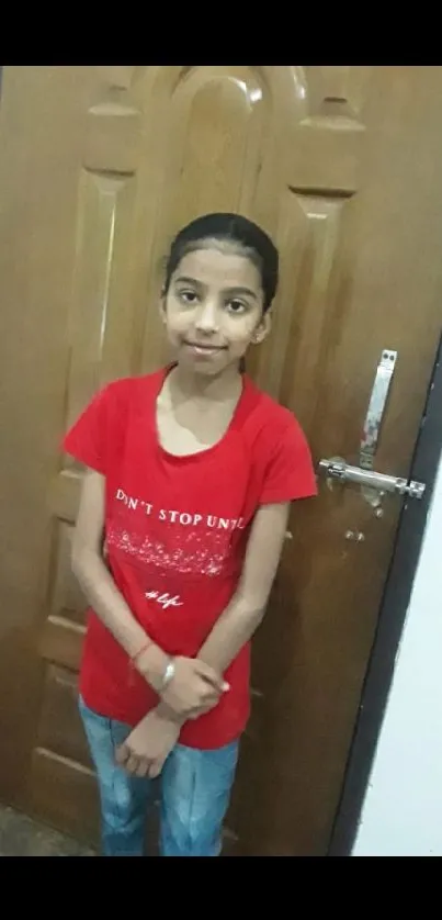 Girl in a red shirt posing in front of a brown wooden door.