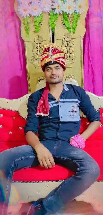 Man in festive attire on red sofa with vibrant backdrop.