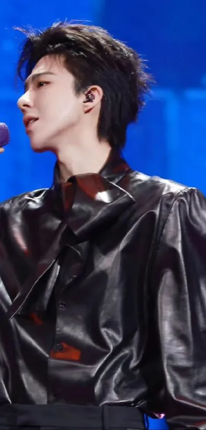 Singer in black outfit with blue background holding a microphone.