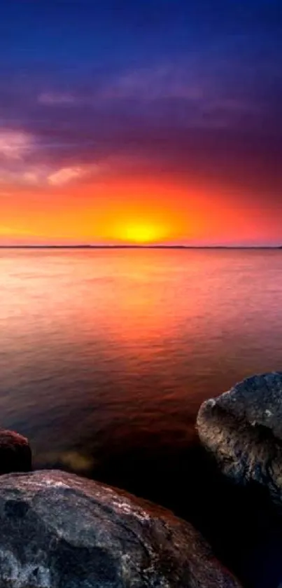 Vibrant sunset over calm waters with rocks in the foreground.