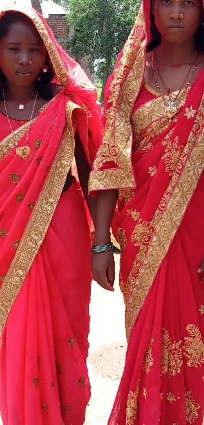 Two women wearing vibrant red sarees with golden embroidery.