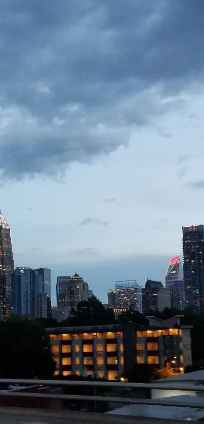 Skyline at dusk with lit skyscrapers under cloudy sky.