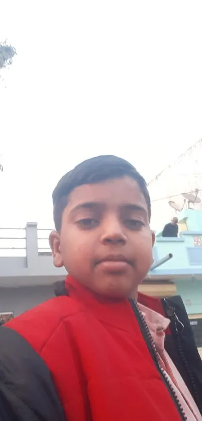 Boy in red jacket on urban street with buildings in background.