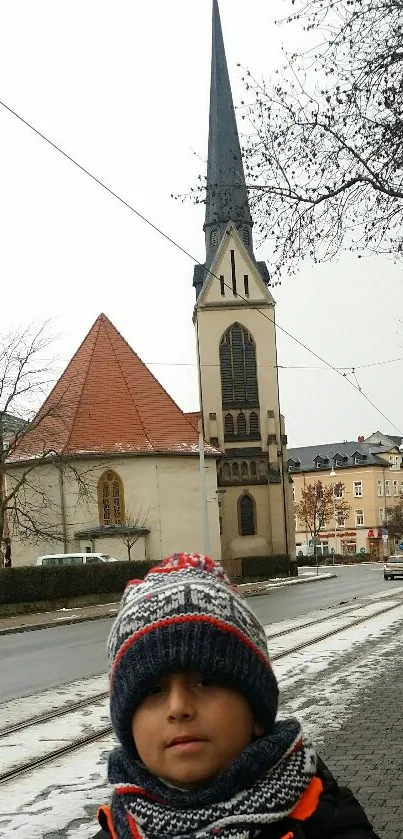 Boy in winter attire stands before a church on a snowy city street.