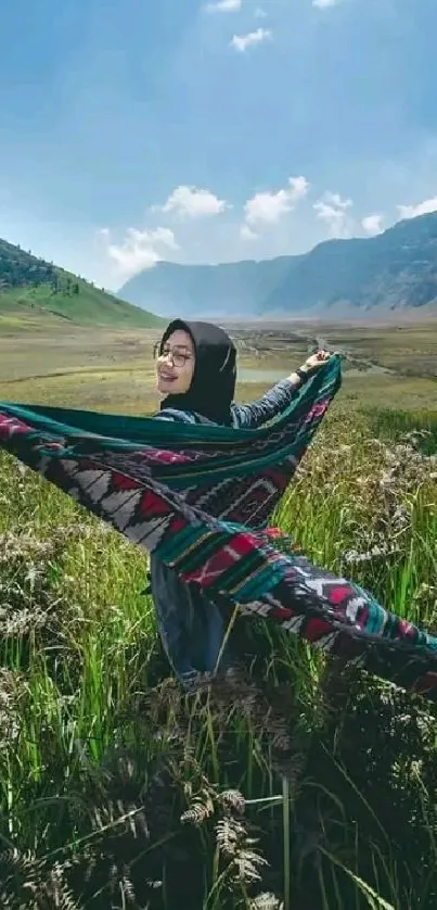 Woman holds a colorful shawl in green meadow with mountains and blue sky.