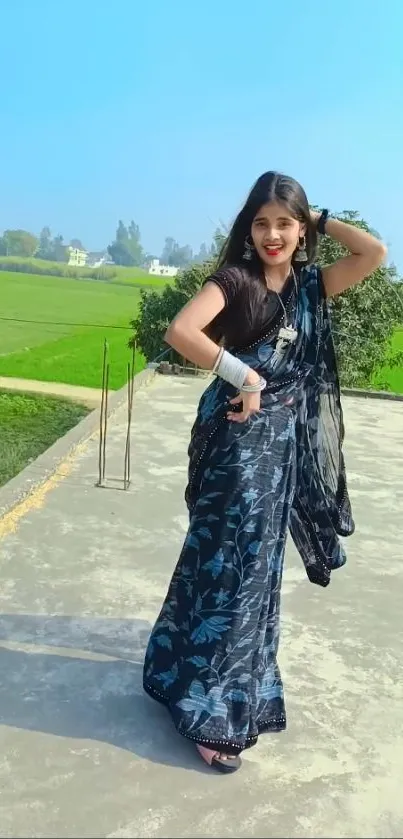 A woman in a vibrant saree poses outdoors against a rural backdrop.