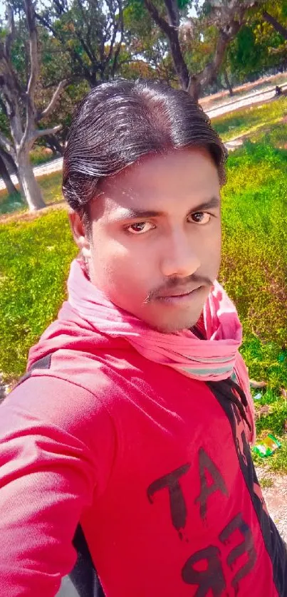 Young man in red shirt outdoors with greenery background.