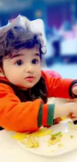 Adorable baby in orange jacket eating on a plate.