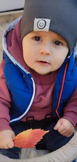 Baby in fall attire holding a leaf, seated outdoors.