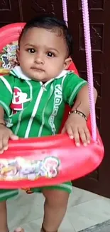 Adorable baby in a green outfit sitting on a red swing.