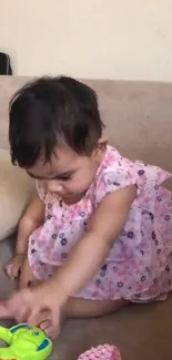 Baby in pink dress plays with toy on beige couch.